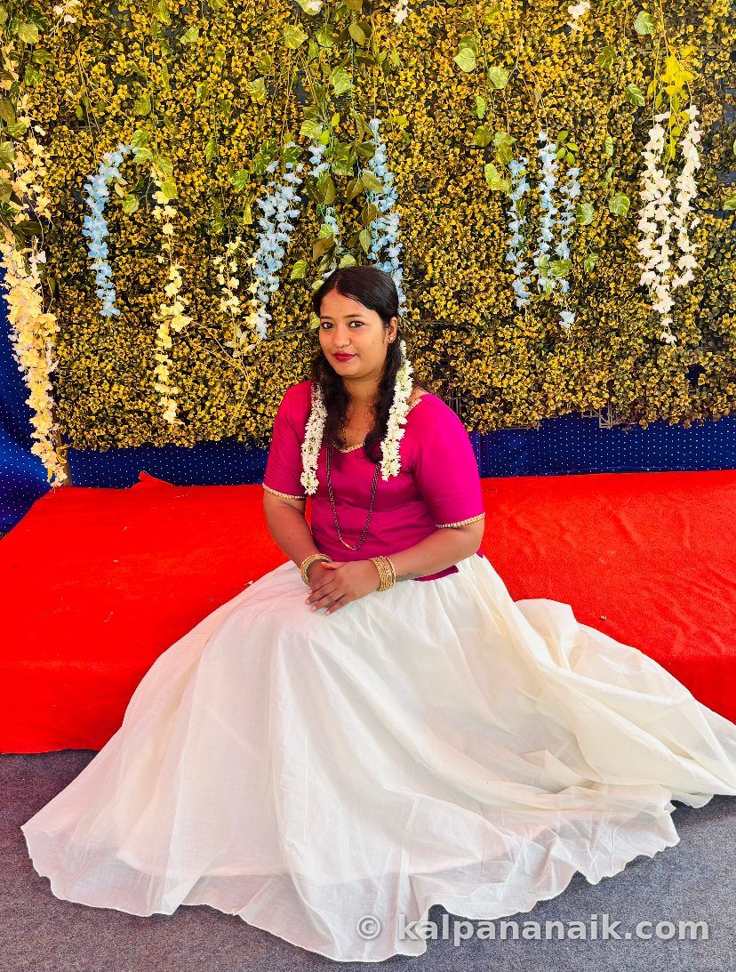 Kalpana in a magenta blouse and white skirt with jasmine in her hair, seated before a flower wall