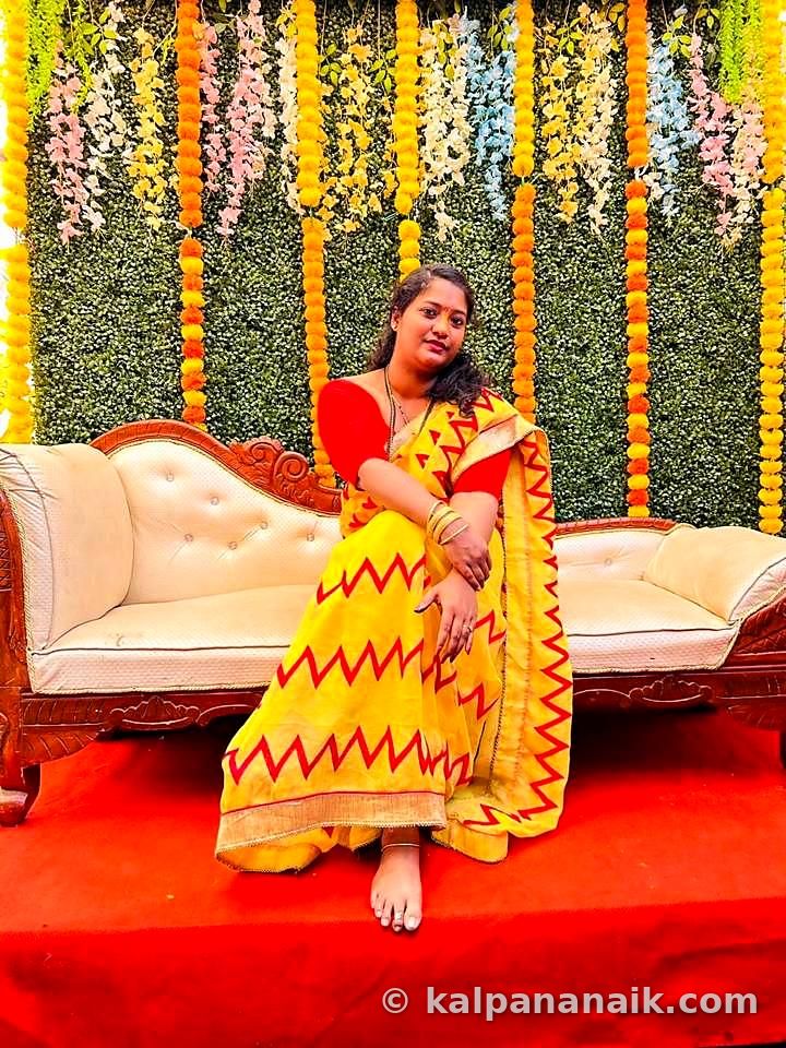 Kalpana in a yellow and red saree seated on a white sofa